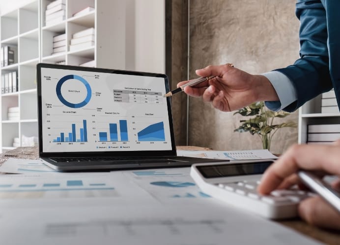 A close-up of a laptop screen displaying various blue financial charts and graphs, including a donut chart and bar graphs. A hand in a blue suit points a pen at the data while another person uses a white calculator in the foreground.