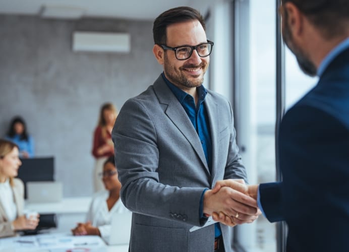 A smiling businessman in a grey suit and glasses shaking hands with a colleague in a bright, modern office setting. Other professionals are visible in the background, suggesting a successful meeting or new partnership.