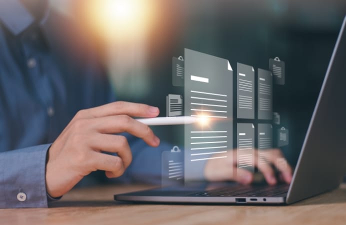 A person's hand using a stylus to select digital document icons that are hovering in front of a laptop screen. The image represents digital file management, cloud storage, and paperless office solutions.
