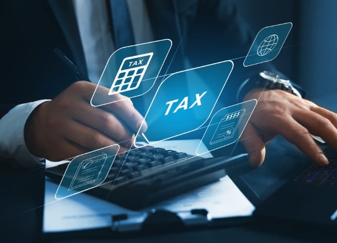 A businessman in a suit using a calculator and pen while working on tax documents. Digital blue icons representing "TAX," a calculator, a globe, and financial reports are superimposed over the scene to represent digital accounting and tax preparation.