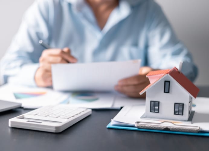 A close-up shot of a small model house with a red roof sitting on paperwork, with a person blurred in the background reviewing documents and using a calculator.