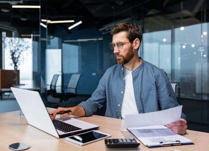A focused male professional with glasses and a beard working at a modern office desk. He is holding a document in one hand and typing on a white laptop with the other, surrounded by a tablet, calculator, and financial charts.